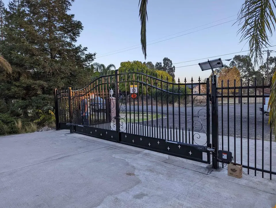 A narrow path lined with sturdy Reno Fence and Gate installations on both sides. The ground is covered with gravel, and a small concrete walkway is visible at the entrance. Houses and greenery are in the background under a clear blue sky.