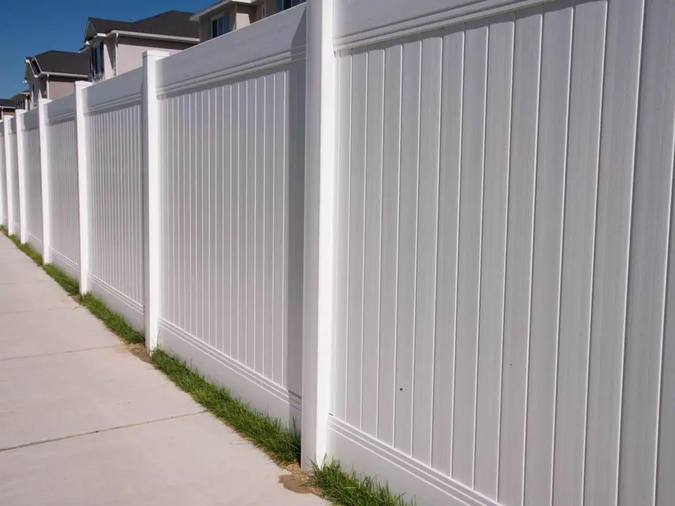 A long white vinyl fence, expertly installed by Reno Fence and Gate, boasts decorative lattice tops running alongside a raised stone-bordered garden. Red flowers bloom in the foreground, with lush green trees visible in the background under a partly cloudy sky.
