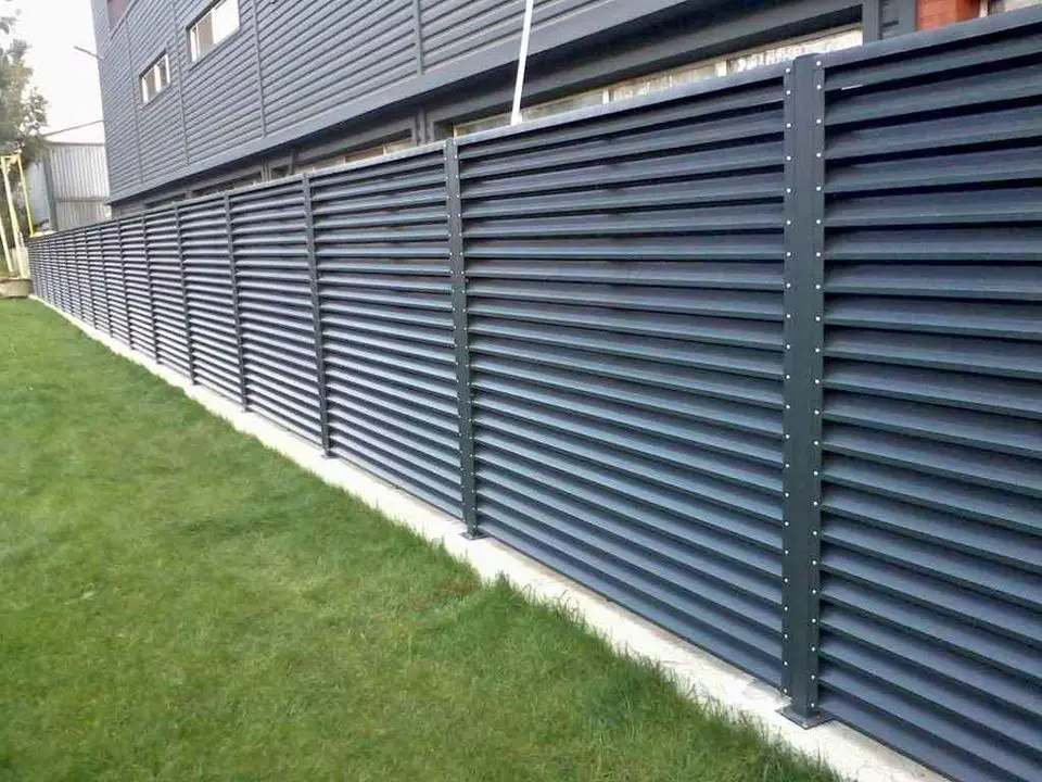A narrow path lined with sturdy Reno Fence and Gate installations on both sides. The ground is covered with gravel, and a small concrete walkway is visible at the entrance. Houses and greenery are in the background under a clear blue sky.