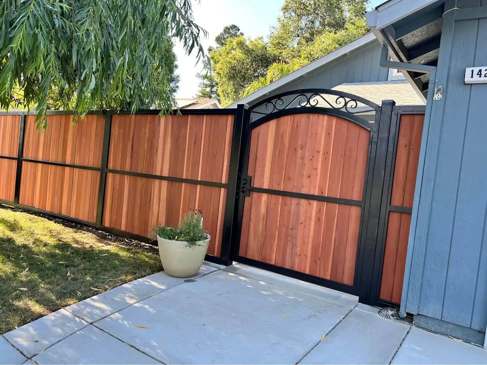 A wooden double gate with black metal framing stands closed in a Reno driveway, nestled between a house and a fence. The sky is clear with some clouds in the background, crafted expertly by a local wood fence contractor renowned for their gate services.
