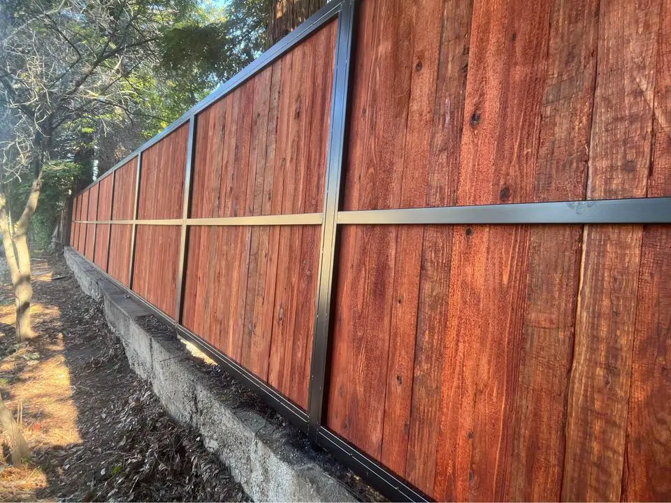 A wooden double gate with black metal framing stands closed in a Reno driveway, nestled between a house and a fence. The sky is clear with some clouds in the background, crafted expertly by a local wood fence contractor renowned for their gate services.