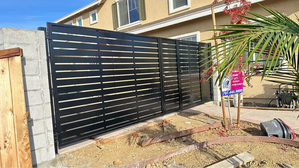 A wooden double gate with black metal framing stands closed in a Reno driveway, nestled between a house and a fence. The sky is clear with some clouds in the background, crafted expertly by a local wood fence contractor renowned for their gate services.