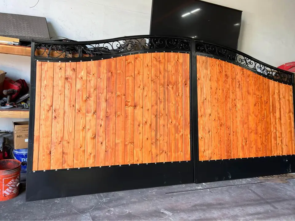 A narrow path lined with sturdy Reno Fence and Gate installations on both sides. The ground is covered with gravel, and a small concrete walkway is visible at the entrance. Houses and greenery are in the background under a clear blue sky.