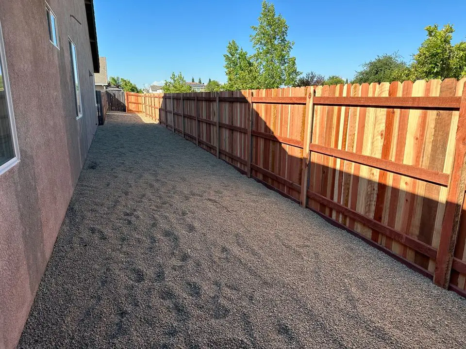 A beige vinyl Reno Fence separates a grassy backyard from a sandy, unfinished area. Gardening tools and equipment are scattered on the ground, with a clear blue sky overhead and neighboring houses visible in the background, hinting at an upcoming gate installation.