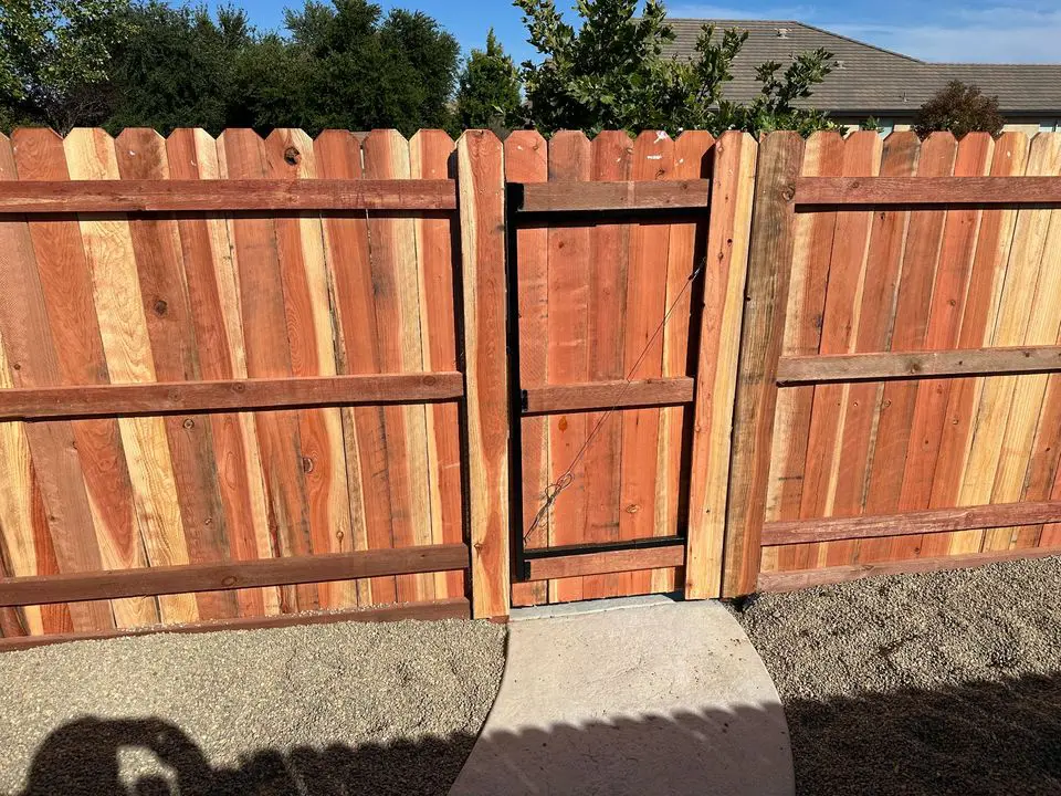 A long white vinyl fence, expertly installed by Reno Fence and Gate, boasts decorative lattice tops running alongside a raised stone-bordered garden. Red flowers bloom in the foreground, with lush green trees visible in the background under a partly cloudy sky.