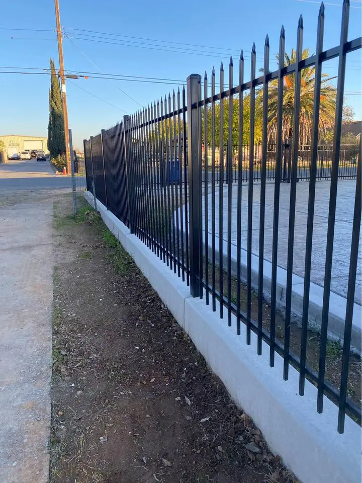 A long, beige vinyl fence crafted by Reno Fence and Gate separates a grassy area from a dirt pathway. Construction tools and materials are scattered along the path. Houses and a clear blue sky are visible in the background, reflecting the quality installation of this sturdy barrier.