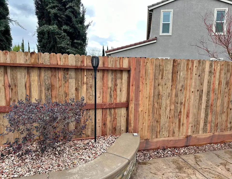 A narrow path lined with sturdy Reno Fence and Gate installations on both sides. The ground is covered with gravel, and a small concrete walkway is visible at the entrance. Houses and greenery are in the background under a clear blue sky.