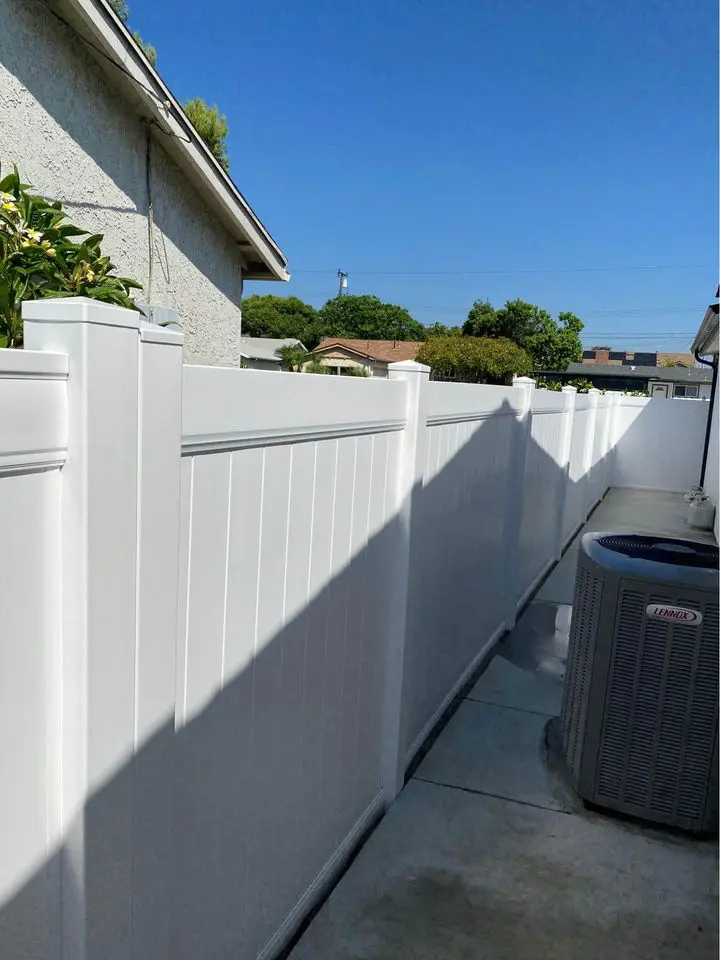 A narrow path lined with sturdy Reno Fence and Gate installations on both sides. The ground is covered with gravel, and a small concrete walkway is visible at the entrance. Houses and greenery are in the background under a clear blue sky.