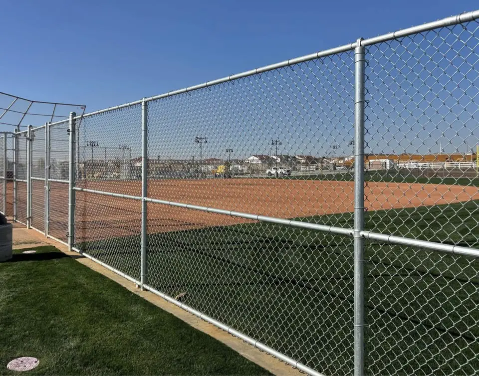A narrow path lined with sturdy Reno Fence and Gate installations on both sides. The ground is covered with gravel, and a small concrete walkway is visible at the entrance. Houses and greenery are in the background under a clear blue sky.